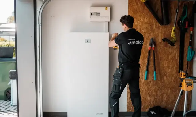 Tekniker installerar ett stort, vitt solpaneler batteri i ett garage. Verktyg hänger på väggen bakom.
