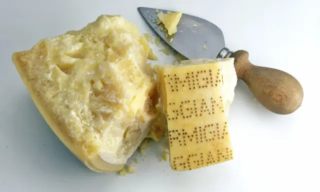 Ein Stück Parmesan mit eingeprägtem Schriftzug und ein Käsemesser. Aus was besteht Parmesan? Aus Milch, Lab und Salz.