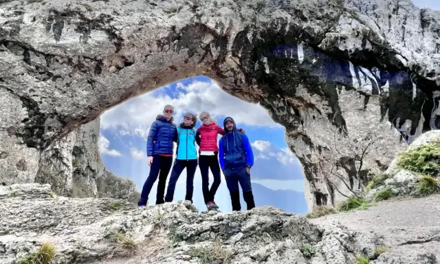 Quattro amici si godono una giornata di camminare in montagna, posando sotto un arco naturale di roccia con un cielo azzurro e nuvoloso.