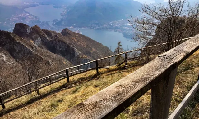 Sentiero con staccionata in legno per una passeggiata sul Pian dei Resinelli, con vista panoramica sul lago e le montagne.