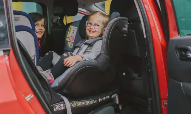 Dwie uśmiechnięte dziewczynki w fotelikach Britax Romer 0-18 kg w czerwonym samochodzie.