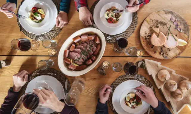 Un repas du terroir avec viande, saucisses, fromages et pain sur une table en bois. Des mains servent et dégustent.