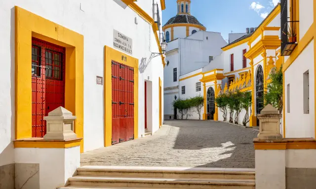 En solig gränd i Sevilla, Spanien, med vita hus, gula detaljer och röda dörrar. En kupol syns i bakgrunden.