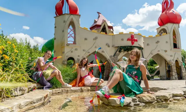 Trzy wróżki w kolorowych strojach siedzą przy strumyku przed bajkowym zamkiem. To z pewnością jedne z wielu magiczne ogrody atrakcje.