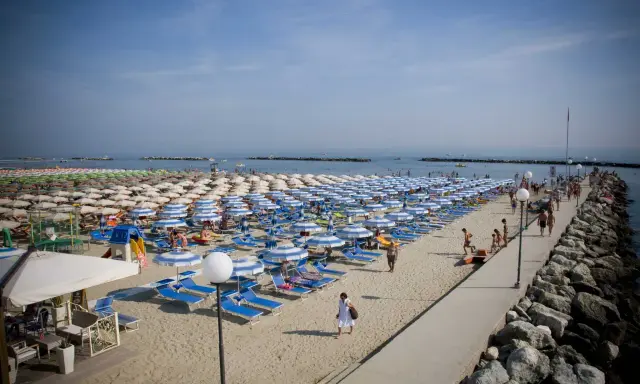 Spiaggia affollata a Rimini Gatteo Mare, con file ordinate di ombrelloni e lettini blu e bianchi, bagnanti e un molo di roccia.