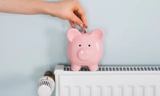 Mano inserta moneda en hucha rosa sobre radiador blanco. Ahorrando energía cuando se apaga la calefacción central.