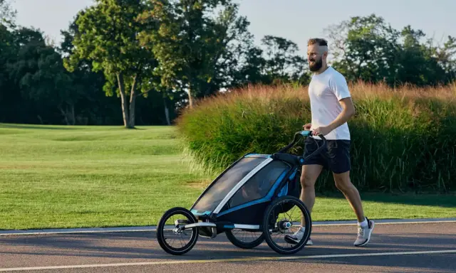 Ein Mann joggt mit Kinderwagen durch eine grüne Parklandschaft.