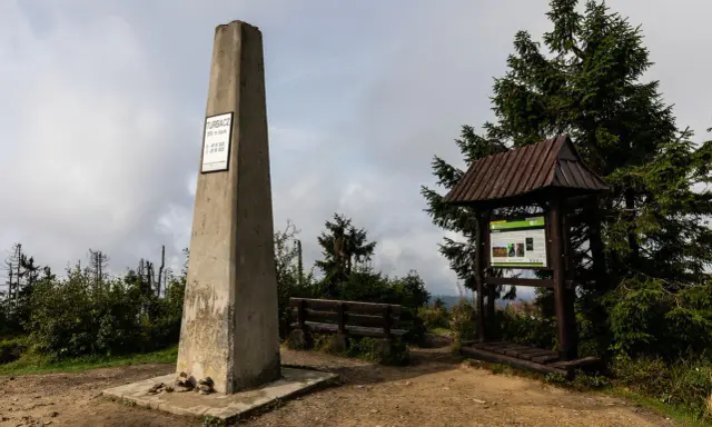 Kamienny obelisk na szczycie Turbacz w Gorcach, obok drewniana wiata z tablicą informacyjną i ławką.