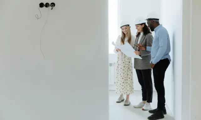 Tres arquitectos con cascos revisan planos en una obra, buscando cómo abaratar costes.