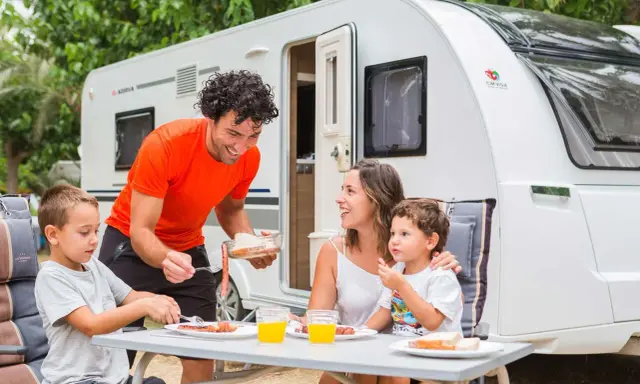 Familia disfrutando de un picnic en campamentos para familias, con una caravana de fondo.