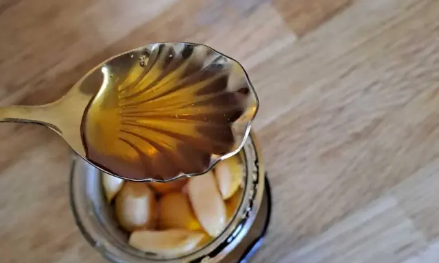 Une cuillère dorée verse du miel ambré sur des gousses d'ail confites dans un bocal. Un délice d'ail au miel.
