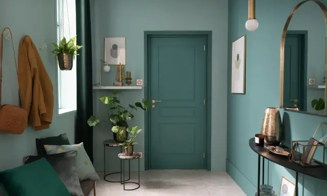 Entrada decorada con pared verde azulado, puerta verde, perchero con abrigo y bolso, plantas y espejo.