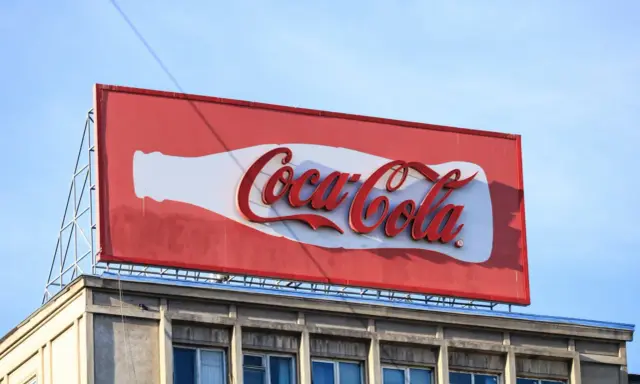 Duży baner logo Coca-Cola na dachu budynku, z charakterystyczną białą butelką i czerwonym napisem.