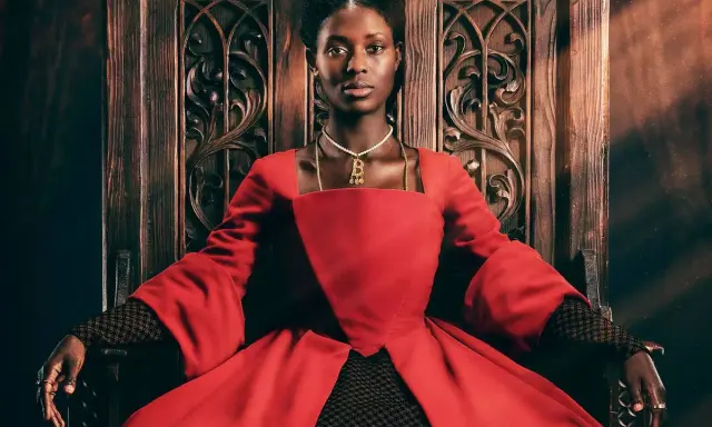 Jodie Turner-Smith, con su mirada regia, se sienta en un trono de madera tallada, vestida con un opulento traje rojo.