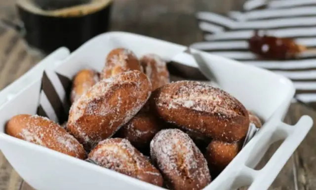 Beignets alsaciens schenkele dorés et saupoudrés de sucre, servis dans un plat blanc avec une tasse de café en arrière-plan.