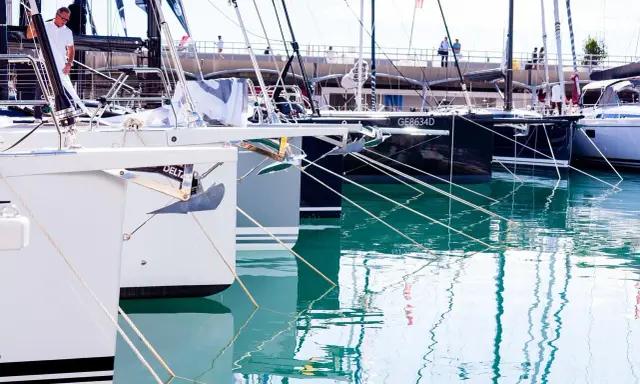 Plusieurs bateaux de plaisance sont amarrés dans un port. La coque du bateau, blanche et grise, est visible au premier plan, avec une ancre argentée.
