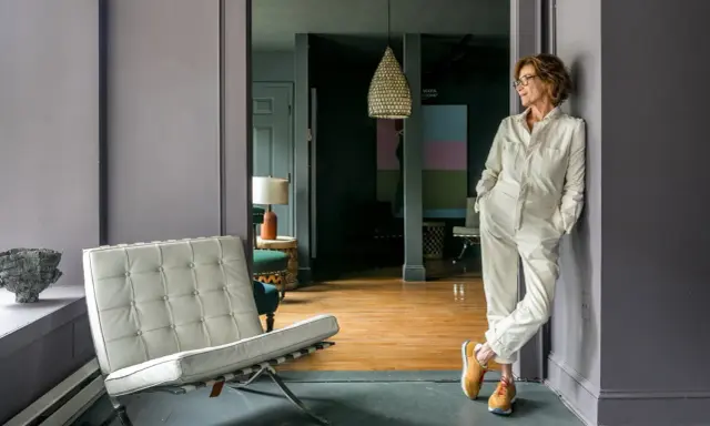 A woman in a cream jumpsuit leans against a wall in the robin rice gallery, with a Barcelona chair and abstract art visible.