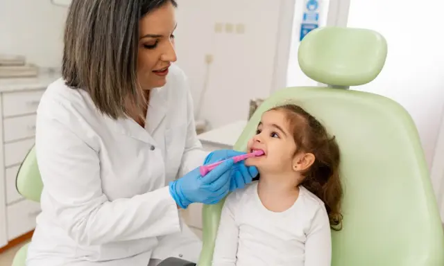 Dentystka w białym fartuchu i niebieskich rękawiczkach pokazuje małej dziewczynce, jak prawidłowo szczotkować zęby. To przykład troski o zdrowie jamy ustnej w ramach pedodoncji.