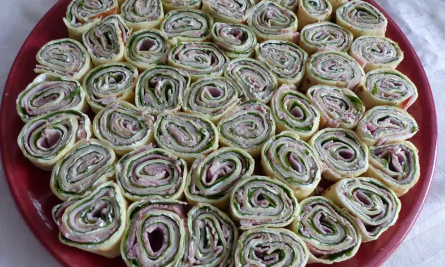 Une assiette rouge remplie de délicieuses crêpes roulées apéritif, garnies de jambon, fromage frais et herbes. Parfait pour un apéritif convivial.