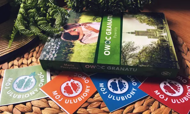 Książka "Owoc granatu" Marii Paszyńskiej z motywem pary na okładce, otoczona zielenią i kolorowymi naklejkami z symbolem granatu.