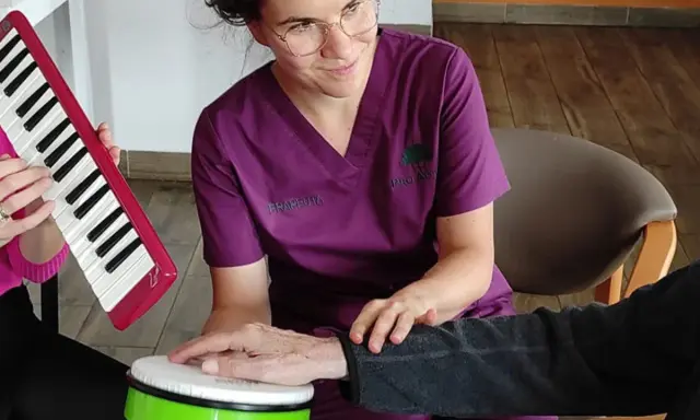 Terapia dźwiękiem i rytmem: muzykoterapia dla seniorów. Terapeuta wspiera dłoń pacjenta na bębenku, podczas gdy inny uczestnik gra na keyboardzie.