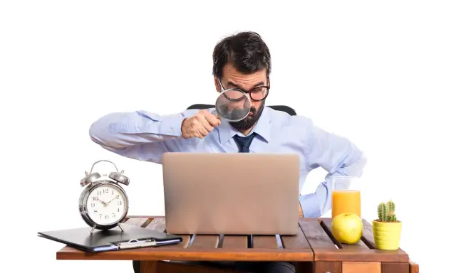 Mężczyzna z lupą bada laptopa, szukając odpowiedzi, czy skstore jest legit. Na biurku budzik, jabłko i sok.
