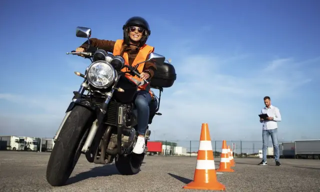 Mujer sonriente en moto, con chaleco naranja y casco, practicando en circuito con conos. Instructor observa. ¡Aprobando el examen de carbet a2!