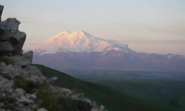 Elbrus, najwyższy szczyt Rosji, lśni w blasku zachodzącego słońca. W oddali widać zielone wzgórza i skaliste zbocze.