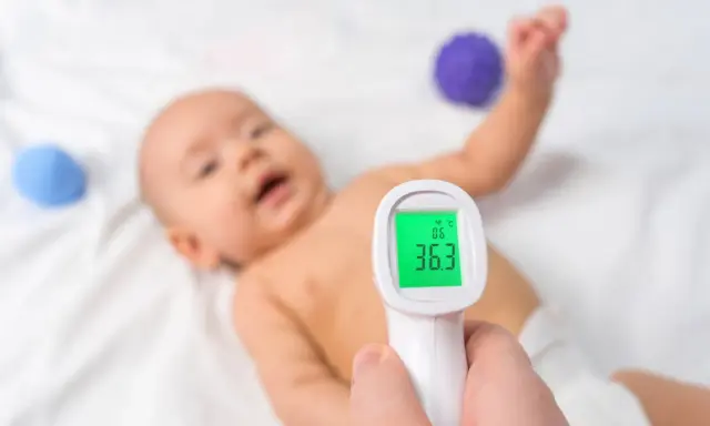 Jak mierzyć temperaturę termometrem elektronicznym? Dziecko leży na białym tle, a termometr pokazuje 36.3°C.