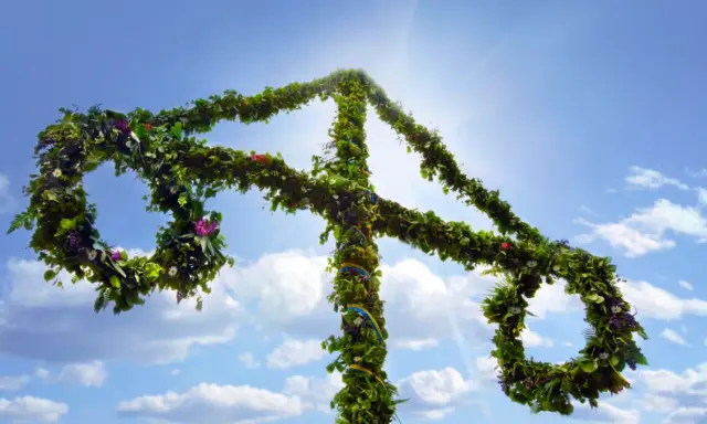 En midsommarstång, klädd i grönska och blommor, reser sig mot en klarblå himmel med solsken.