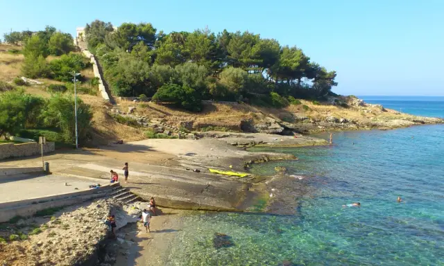 Scogliera e mare cristallino a San Gregorio Leuca. Persone si godono il sole e l'acqua, una scalinata sale verso la collina alberata.