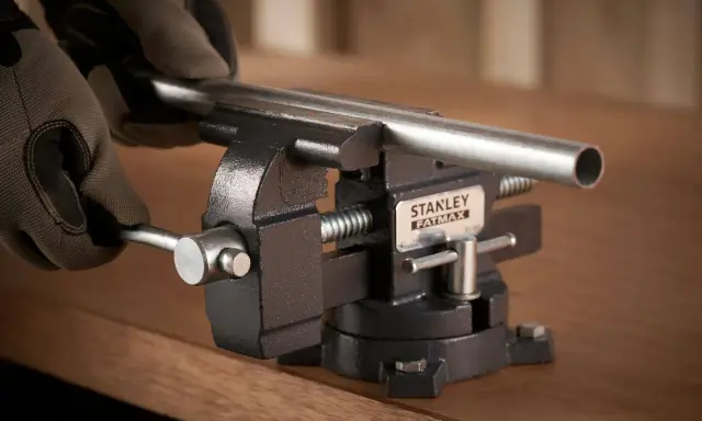 Manos enguantadas ajustan un tubo metálico en un tornillo de banco Stanley Fatmax, herramienta esencial para fijar piezas.