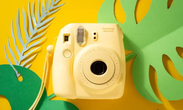 Żółty aparat natychmiastowy Fujifilm Instax mini 8 na tle tropikalnych liści.
