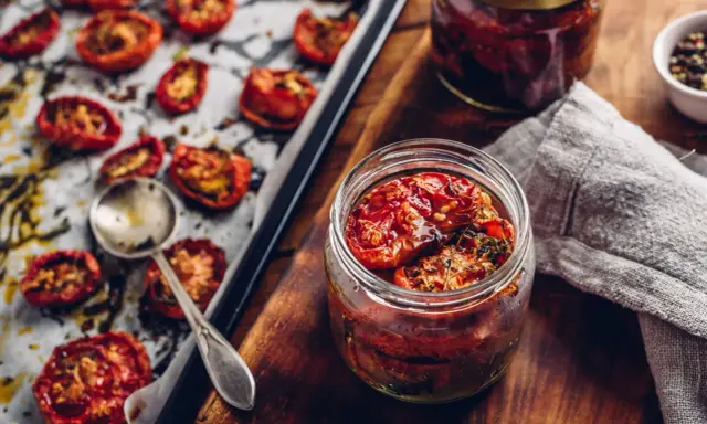 Pomidory suszone w oleju z ziołami przepis. Słoik pełen aromatycznych, suszonych pomidorów w oliwie, obok blachy z pieczonymi pomidorami.