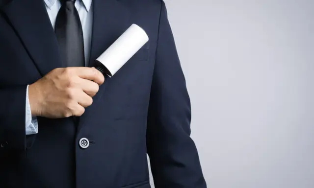 A man in a dark tuxedo uses a lint roller to clean his jacket, showing a simple way how to clean a tuxedo.