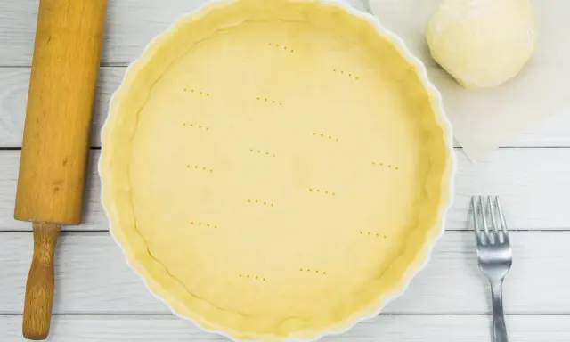 Une pâte brisée est prête dans une casserole blanche, avec une boule de pâte, une rouleau et une fourchette à côté.