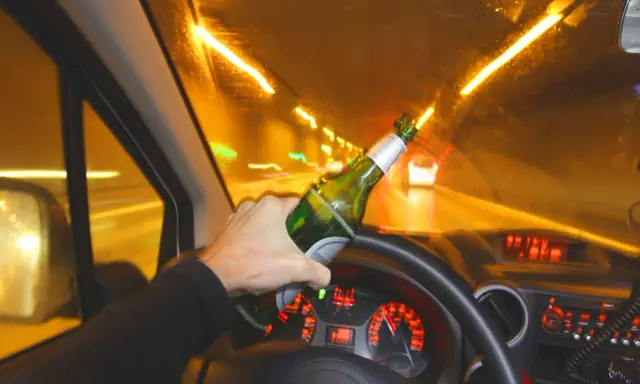 Ręka trzyma butelkę piwa za kierownicą. Po ilu można jechać autem po alkoholu? Nigdy!