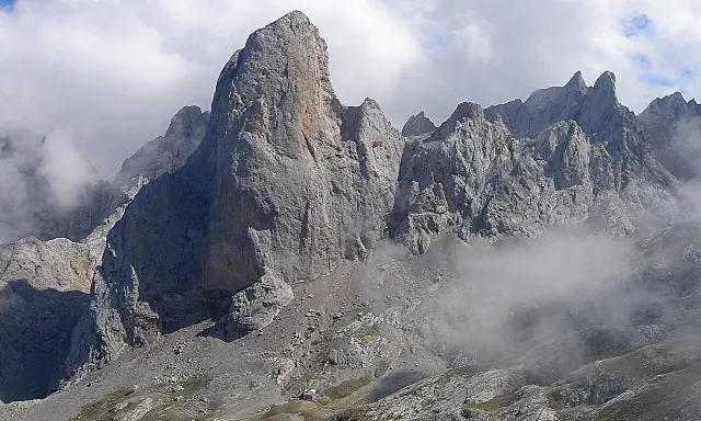 Majestatyczny **Naranjo de Bulnes** w Picos de Europa, otoczony mgłą i chmurami, z małym schroniskiem u podnóża.