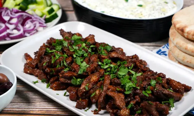 En tallrik med saftigt gyroskött, toppat med färsk persilja. Bredvid syns pitabröd, tzatziki, oliver, gurka och rödlök. Ett perfekt gyros recept!