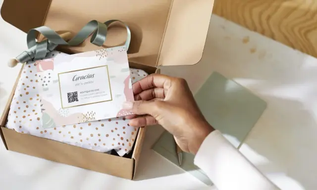 Tarjeta de agradecimiento tamaño postal con código QR, dentro de una caja de regalo abierta con papel de seda dorado.