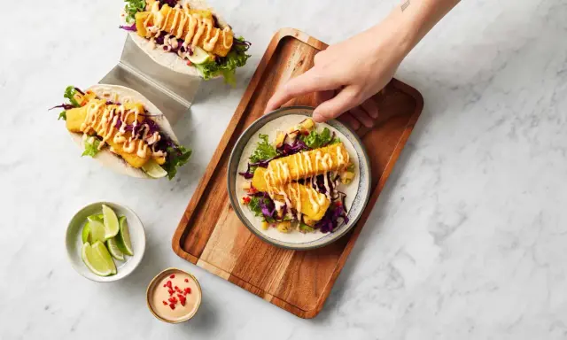 Två läckra fish tacos med krispig fisk, rödkål, sallad och en krämig sås. En hand sträcker sig efter en tallrik med en taco.