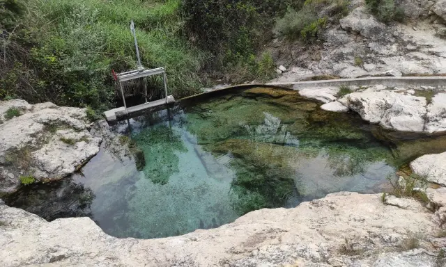 Aguas cristalinas en los baños de Somogil, rodeadas de rocas y vegetación. Una compuerta metálica controla el flujo del agua.