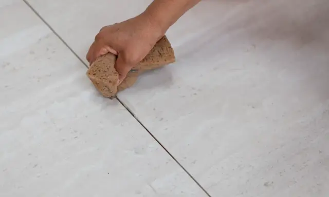 Main d'une personne nettoyant le joint de carrelage avec une éponge. La couleur du joint est sombre, contrastant avec le carrelage clair.