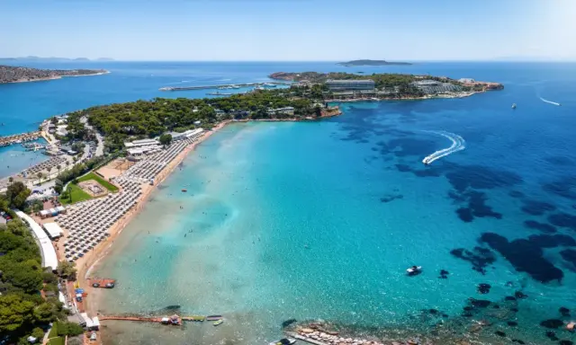 Traumhafter Blick auf das athen meer mit einer belebten Strandpromenade, Liegestühlen und dem glitzernden, türkisfarbenen Wasser, auf dem Boote fahren.