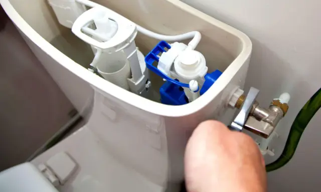 Mécanisme de chasse d'eau qui coule, avec un flotteur bleu et blanc visible. Une main ajuste le robinet d'arrivée d'eau.