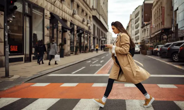 Kobieta w beżowym płaszczu i żółtych sneakersach przechodzi przez pasy. Idealne buty do trencza na miejski spacer.
