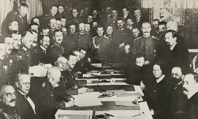 Delegierte unterzeichnen den Frieden von Brest-Litowsk. Männer in Uniformen und Anzügen sitzen an einem Tisch, umgeben von weiteren Personen.