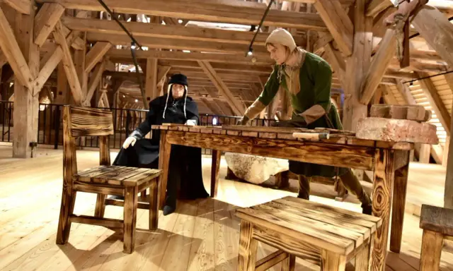 W muzeum Mazury dwie figury w strojach historycznych przy drewnianym stole. Jedna siedzi, druga stoi, pracując.