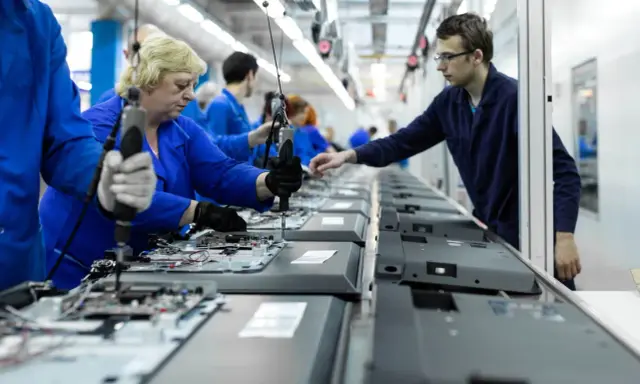Pracownicy w niebieskich kombinezonach montują telewizory na linii produkcyjnej. To praca w ruchu ciągłym, gdzie każdy element jest kluczowy.