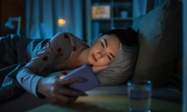 Une jeune femme dans son lit, le visage éclairé par son téléphone. Trop d'écrans, conséquences sur le sommeil.